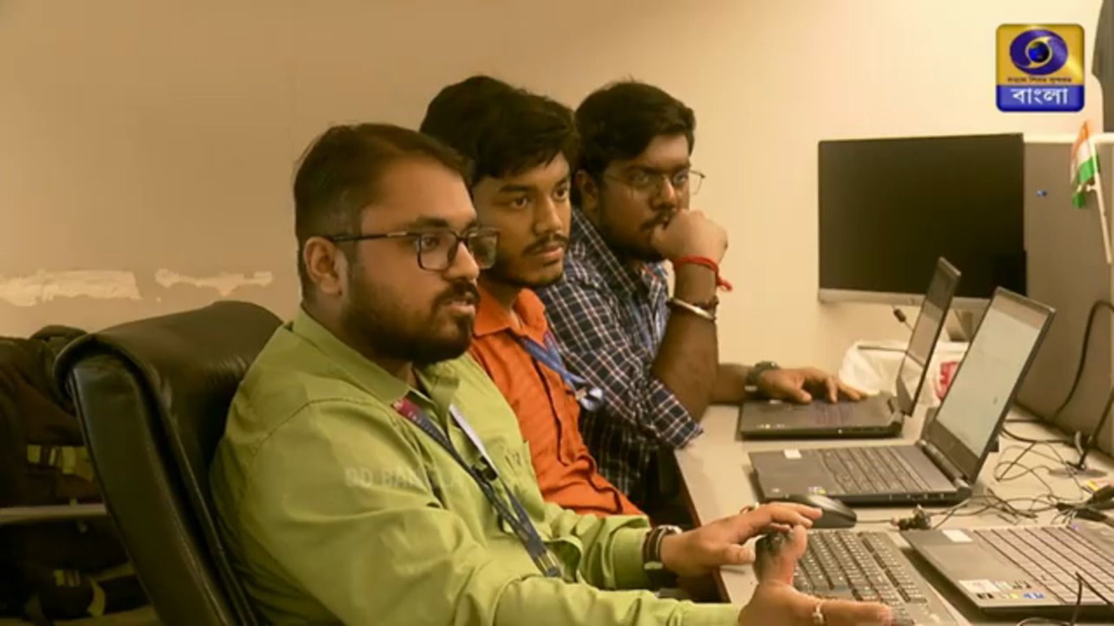 Wide still of the office desk: three colleagues at laptops, with a desktop monitor, cabling and the Indian flag visible behind them.